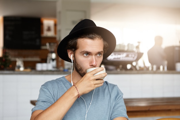 Tiro al coperto di giovane attraente in cappello alla moda che beve caffè o tè, rilassante al bar da solo, ascoltando musica online sugli auricolari