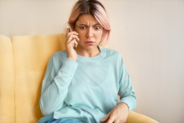 Tiro al coperto di giovane donna caucasica emotiva che indossa anello al naso e felpa blu che riceve cattive notizie mentre parla al telefono cellulare, avendo spaventato l'espressione del viso scioccato, sopracciglia accigliate