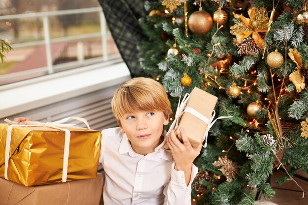 Tiro al coperto di ragazzo adolescente biondo seduto sotto l'albero di Capodanno decorato circondato da regali di Natale, scatola tremante, cercando di indovinare cosa c'è dentro, con un'espressione facciale curiosa e interessata