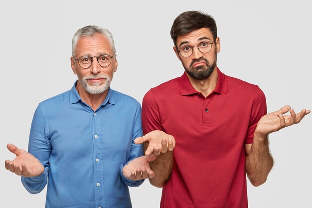 Tiro al coperto di un maschio maturo perplesso e suo figlio adulto, alzando le spalle, non possono prendere una decisione, essendo con la barba lunga, stanno vicini l'uno all'altro contro il muro bianco. Allora cosa fare in questa situazione?