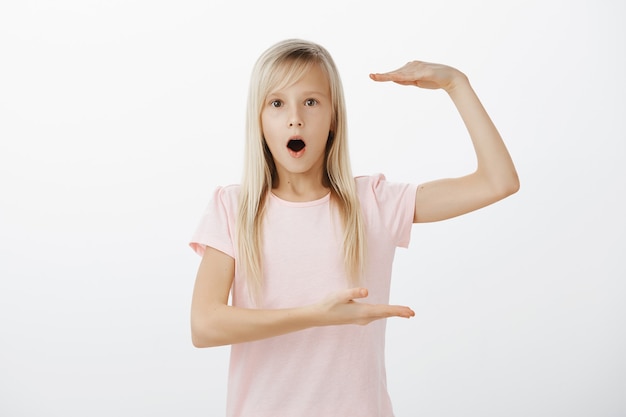 Tiro al coperto di una bambina adorabile scioccata con i capelli biondi, mascella cadente e forma di qualcosa di grande con due palme, stupita dalle dimensioni dell'oggetto sul muro grigio