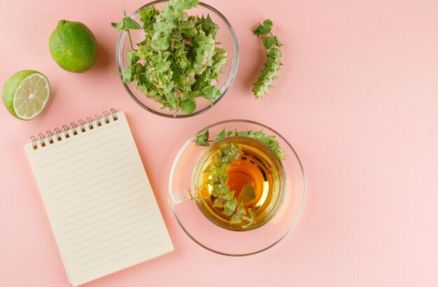 Tisana con erbe, lime, taccuino in una tazza di vetro su rosa, vista dall'alto.