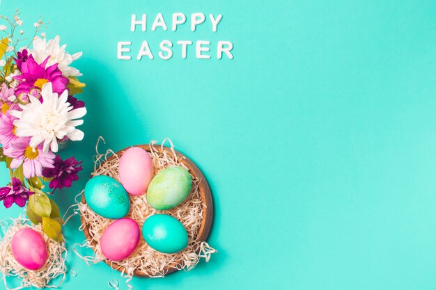 Titolo di Pasqua felice vicino a uova luminose sul piatto e bouquet di fiori