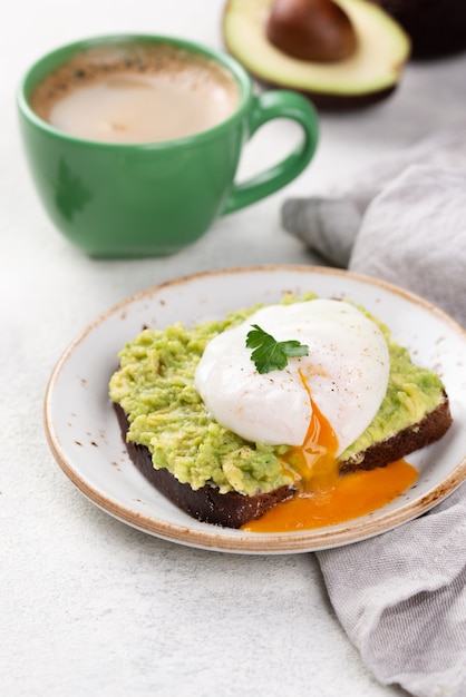 Toast di avocado sul piatto con uovo affogato che cola in cima e tazza di caffè