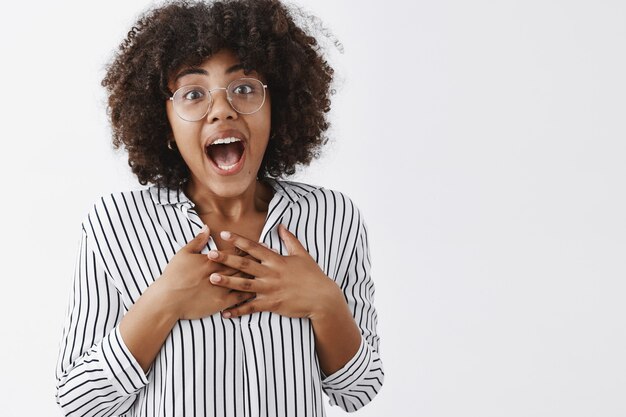 toccato sorpreso e grato attraente giovane collega femminile con la pelle scura, acconciatura afro in camicetta a righe tenendo le mani sul seno e sorridente con la bocca aperta dallo stupore