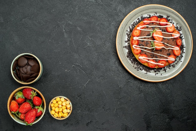 Torta appetitosa vista dall'alto con pezzi di cioccolato e fragola sul lato destro del tavolo e ciotole di cioccolato fragola e nocciola sul lato sinistro del tavolo scuro