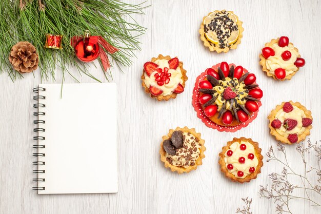 Torta di bacche vista dall'alto arrotondata con foglie di albero di pino crostate con giocattoli di Natale e un taccuino sul terreno in legno bianco