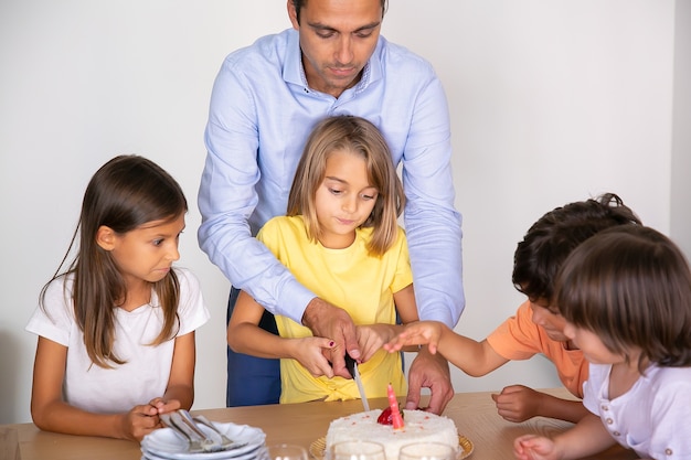 Torta di compleanno di taglio della bambina sveglia con l'aiuto del padre. Bambini adorabili felici che festeggiano il compleanno insieme e aspettano il dessert in sala da pranzo. Infanzia, celebrazione e concetto di vacanza