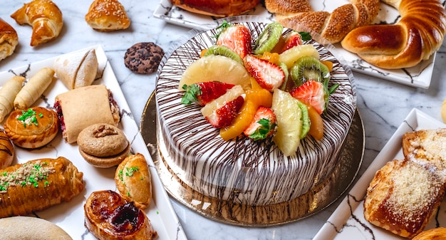 Torta di frutta vista laterale con crema di vaniglia cioccolato kiwi arancione fragola ananas e pasticcini sul tavolo