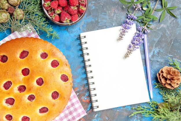 Torta di lamponi a metà vista dall'alto sulla ciotola dell'asciugamano da cucina con lamponi ramo di pino un quaderno una penna su sfondo blu