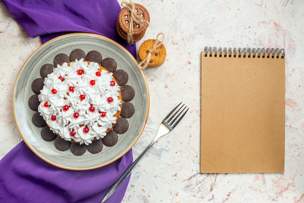 Torta vista dall'alto con crema pasticcera su piatto ovale biscotti a scialle viola legati con taccuino a forchetta in corda
