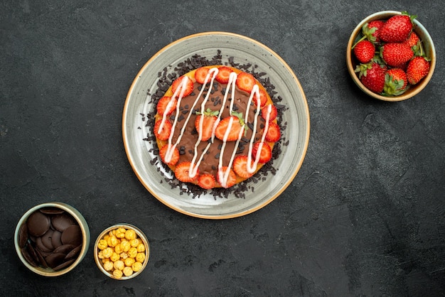 Torta vista dall'alto con piatto di torta al cioccolato con cioccolato e fragola tra ciotole di nocciola fragola e cioccolato sul tavolo scuro