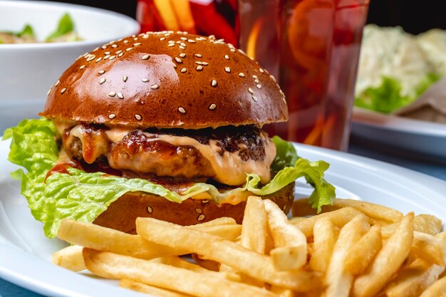 Tortino di manzo grigliato hamburger di vista laterale con lattuga di salsa di formaggio fuso tra panini hamburger e patatine fritte sul tavolo