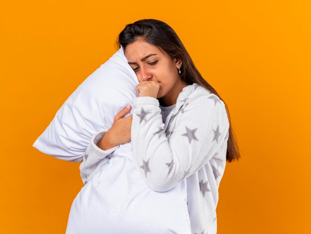 Tosse giovane ragazza malata guardando verso il basso abbracciando cuscino mettendo la mano sulla bocca isolata su giallo