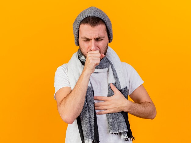 Tosse giovane uomo malato che indossa cappello invernale e sciarpa mettendo la mano sul petto isolato su giallo