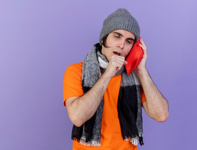 Tosse giovane uomo malato che indossa un cappello invernale con sciarpa che mette il sacchetto di acqua calda sulla guancia isolato su sfondo viola