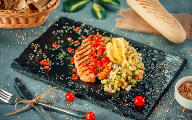 Trancio di salmone con insalata di limone e verdure su un piatto di pietra nera.