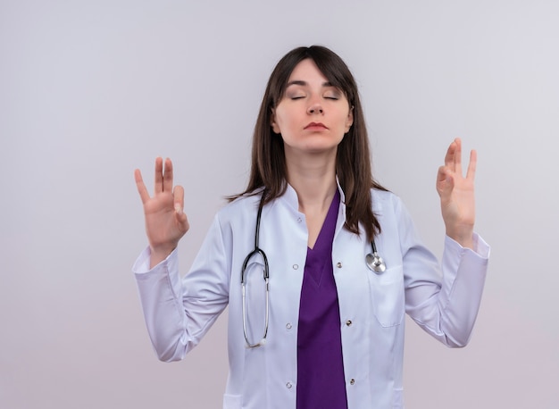 Tranquillo giovane medico femminile in abito medico con lo stetoscopio finge di meditare su sfondo bianco isolato con copia spazio
