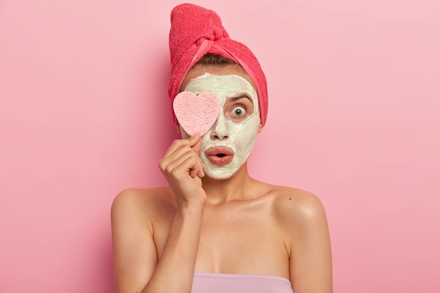 Trattamento viso e concetto spa. La giovane donna stupita applica una maschera all'argilla, scioccata dal risultato rapido ed efficace, mantiene la spugna cosmetica sugli occhi, sta nuda contro il muro rosa, guarisce la pelle