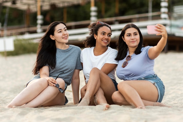 Tre amiche che si siedono sulla spiaggia e prendendo selfie