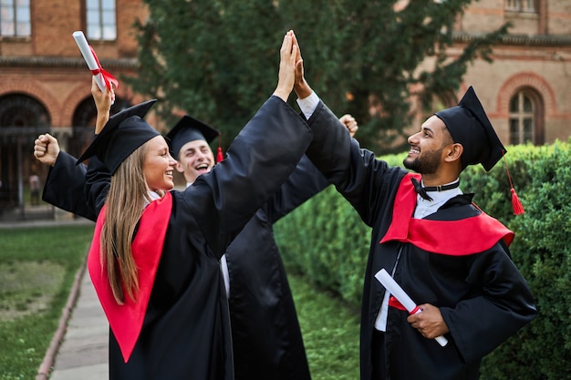 Tre amici laureati internazionali felici che salutano nel campus universitario in abiti di laurea con diploma.