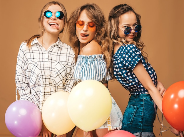 Tre belle donne sorridenti in abiti a scacchi estate camicia. Ragazze in occhiali da sole in posa. Modelli con palloncini colorati. Divertirsi, mostrando la loro lingua