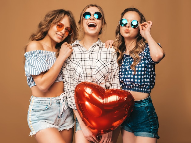 Tre belle donne sorridenti in abiti a scacchi estate camicia. Ragazze in posa Modelli con palloncino a forma di cuore in occhiali da sole. Pronto per la festa di San Valentino