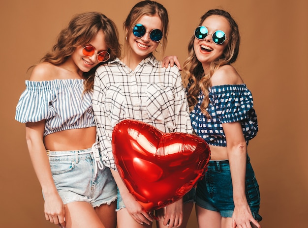 Tre belle donne sorridenti in abiti a scacchi estate camicia. Ragazze in posa Modelli con palloncino a forma di cuore rosso in occhiali da sole. Pronto per la festa di San Valentino