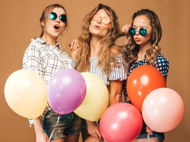 Tre belle donne sorridenti in vestiti a quadretti ed occhiali da sole di estate della camicia. Ragazze in posa Modelli con palloncini colorati. Divertirsi, pronti per la festa di compleanno