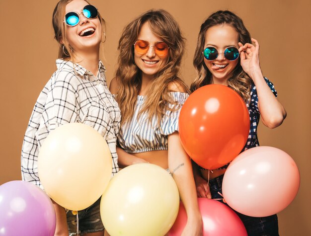 Tre belle donne sorridenti in vestiti a quadretti ed occhiali da sole di estate della camicia. Ragazze in posa Modelli con palloncini colorati. Divertirsi, pronti per la festa di compleanno