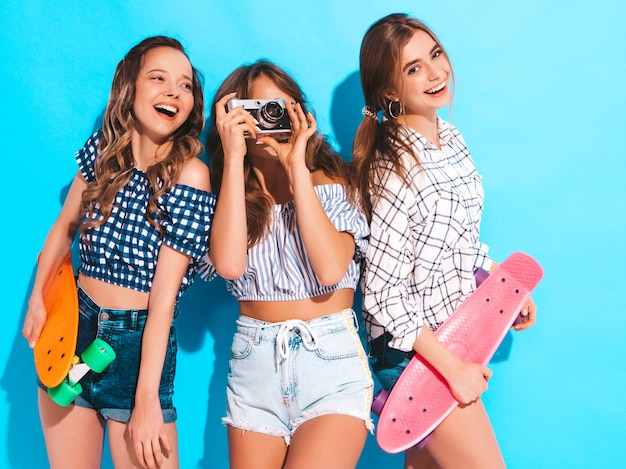 Tre belle ragazze sorridenti alla moda con skateboard penny colorati. Le donne in abiti camicia estiva a scacchi. Scattare foto con la retro macchina fotografica