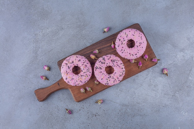 Tre ciambelle dolci rosa con rose in erba sul tagliere di legno.