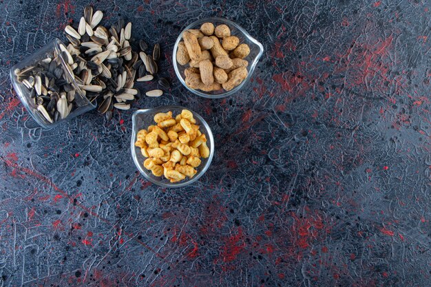 Tre ciotole di arachidi, semi di girasole e cracker su sfondo blu.