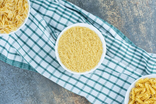 Tre ciotole di pasta accanto a due di grano sull'asciugamano, sullo sfondo di marmo.