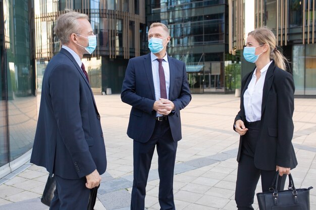 Tre colleghi di lavoro professionisti in maschere facciali discutendo affare al di fuori. Contenuti manager di successo in piedi per strada e parlando di lavoro. Negoziazione, protezione e concetto di partenariato