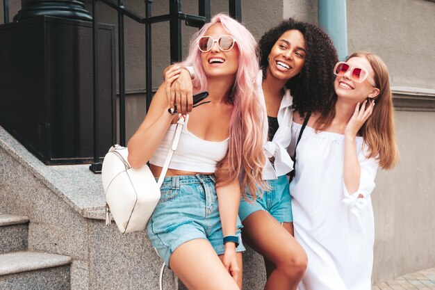 Tre giovani belle donne hipster sorridenti in abiti estivi alla modaDonne multirazziali spensierate sexy in posa sullo sfondo della stradaModelli positivi che si divertono con gli occhiali da sole Allegri e felici