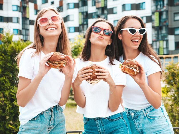 Tre giovani belle donne hipster sorridenti in estate alla moda stessi vestiti Donne sexy spensierate in posa per la stradaModelli positivi che si divertono con gli occhiali da sole Tenendo in mano succosi hamburger e mangiando hamburger