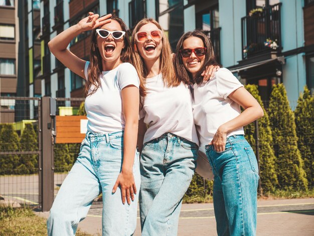 Tre giovani belle donne hipster sorridenti in estate alla moda stessi vestiti Donne sexy spensierate in posa sullo sfondo della stradaModelli positivi che si divertono in occhiali da soleAbbracciare Allegro e felice