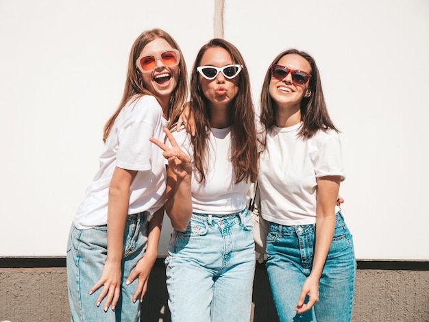 Tre giovani belle donne hipster sorridenti in estate alla moda stessi vestiti Donne sexy spensierate in posa sullo sfondo della stradaModelli positivi che si divertono in occhiali da soleAbbracciare Allegro e felice
