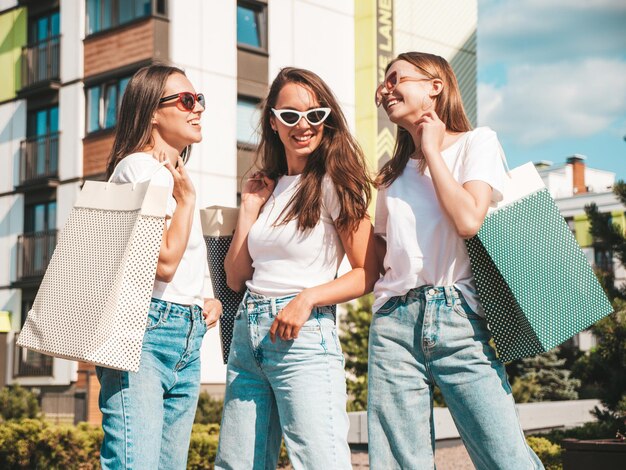 Tre giovani belle donne hipster sorridenti in estate alla moda stessi vestiti Donne sexy spensierate in posa sullo sfondo della stradaModelli positivi che si divertono in occhiali da soleAbbracciare Tenendo le borse della spesa