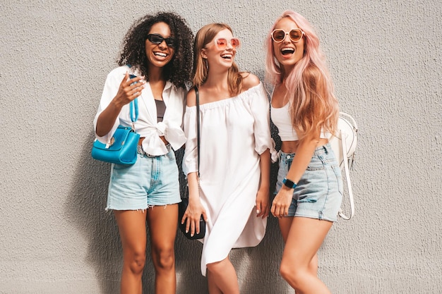 Tre giovani belle donne sorridenti in abiti estivi alla modaDonne multirazziali spensierate sexy in posa sullo sfondo della stradaModelli positivi che si divertono con gli occhiali da sole Allegri e felici