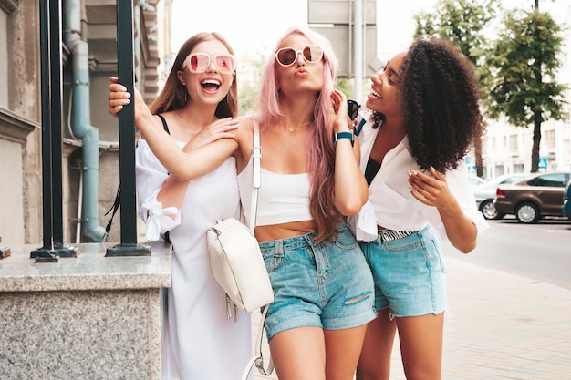 Tre giovani belle donne sorridenti in abiti estivi alla modaDonne multirazziali spensierate sexy in posa sullo sfondo della stradaModelli positivi che si divertono con gli occhiali da sole Allegri e felici