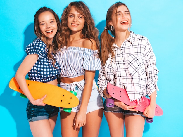 Tre giovani belle ragazze sorridenti alla moda con i pattini variopinti del penny. Donne in estate camicia a scacchi vestiti in posa. Modelli positivi che si divertono