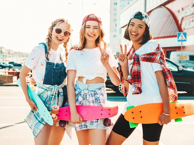 Tre giovani belle ragazze sorridenti con colorati penny skateboard. Donne in abiti estivi hipster in posa sullo sfondo di strada. Modelli positivi che si divertono e impazziscono. Mostra il segno di pace