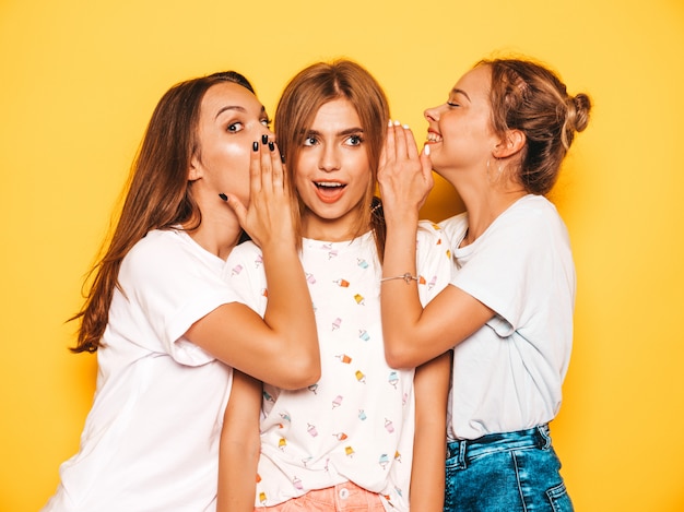 Tre giovani belle ragazze sorridenti hipster in abiti estivi alla moda. Donne spensierate sexy che posano vicino alla parete gialla. Modelle positive che impazziscono e si divertono. Condividi segreti, pettegolezzi