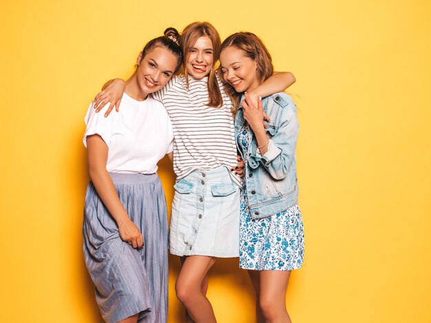 Tre giovani belle ragazze sorridenti hipster in abiti estivi alla moda. Donne spensierate sexy che posano vicino alla parete gialla. Modelli positivi che si divertono