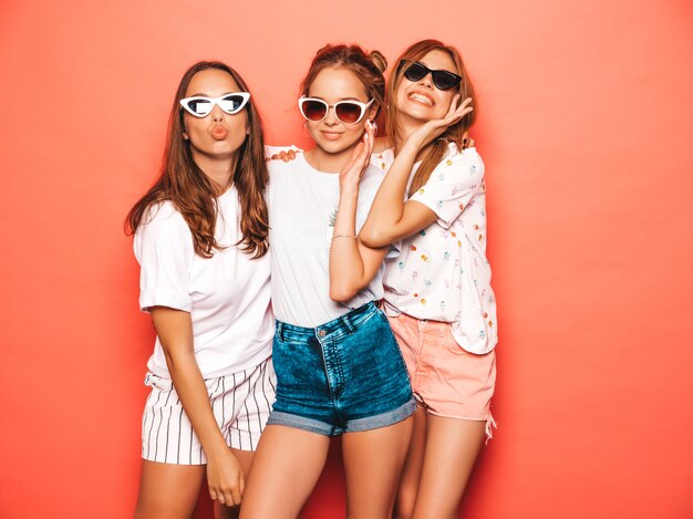Tre giovani belle ragazze sorridenti hipster in abiti estivi alla moda. Donne spensierate sexy che posano vicino alla parete rosa. Modelle positive che impazziscono e si divertono