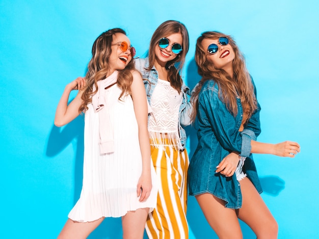 Tre giovani belle ragazze sorridenti in abiti colorati estate alla moda. Donne spensierate sexy in occhiali da sole isolati sul blu. Modelli positivi