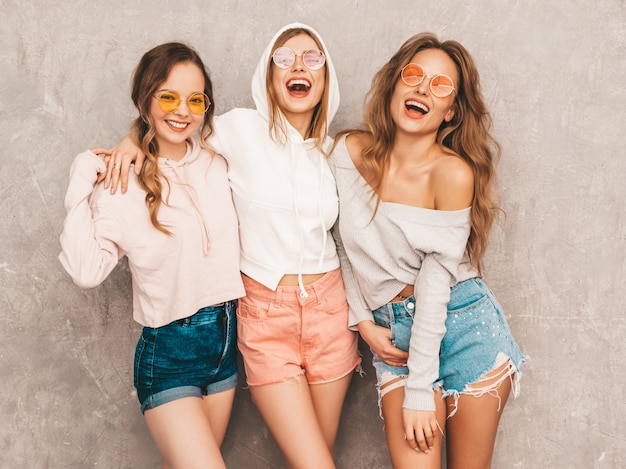 Tre giovani belle ragazze sorridenti in abiti sportivi alla moda estate. Posa sexy spensierata delle donne. Modelli positivi in occhiali da sole rotondi che si divertono. Abbracciare