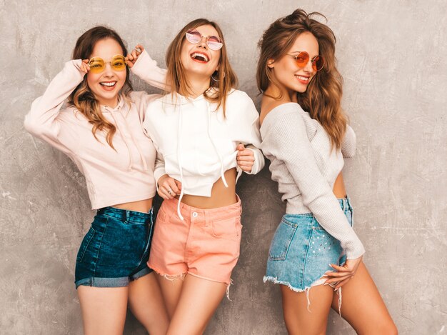 Tre giovani belle ragazze sorridenti in abiti sportivi alla moda estate. Posa sexy spensierata delle donne. Modelli positivi in occhiali da sole rotondi che si divertono. Abbracciare